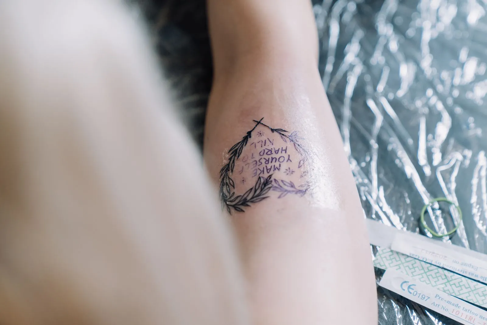 Close-up of a fresh tattoo with inspirational text encircled by laurel wreath on skin.